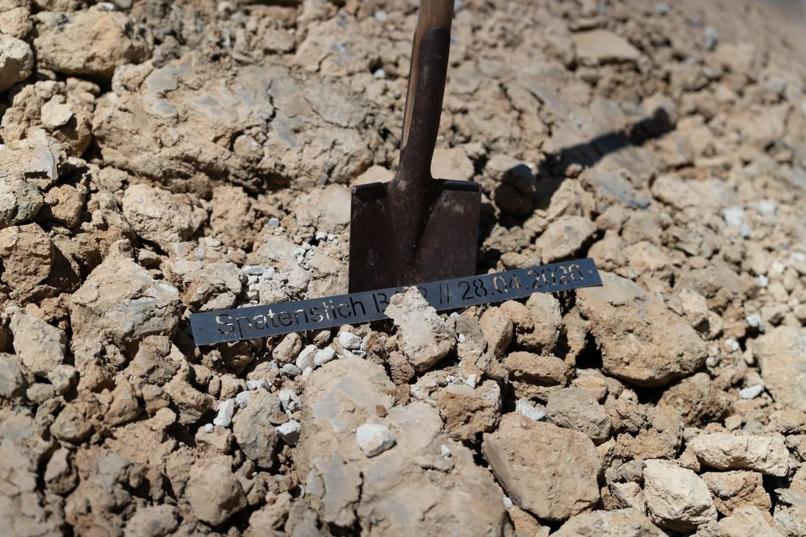 Metalen bord „Eerste spade BA 2 // 28.04.2026“ in de aarde naast een spade